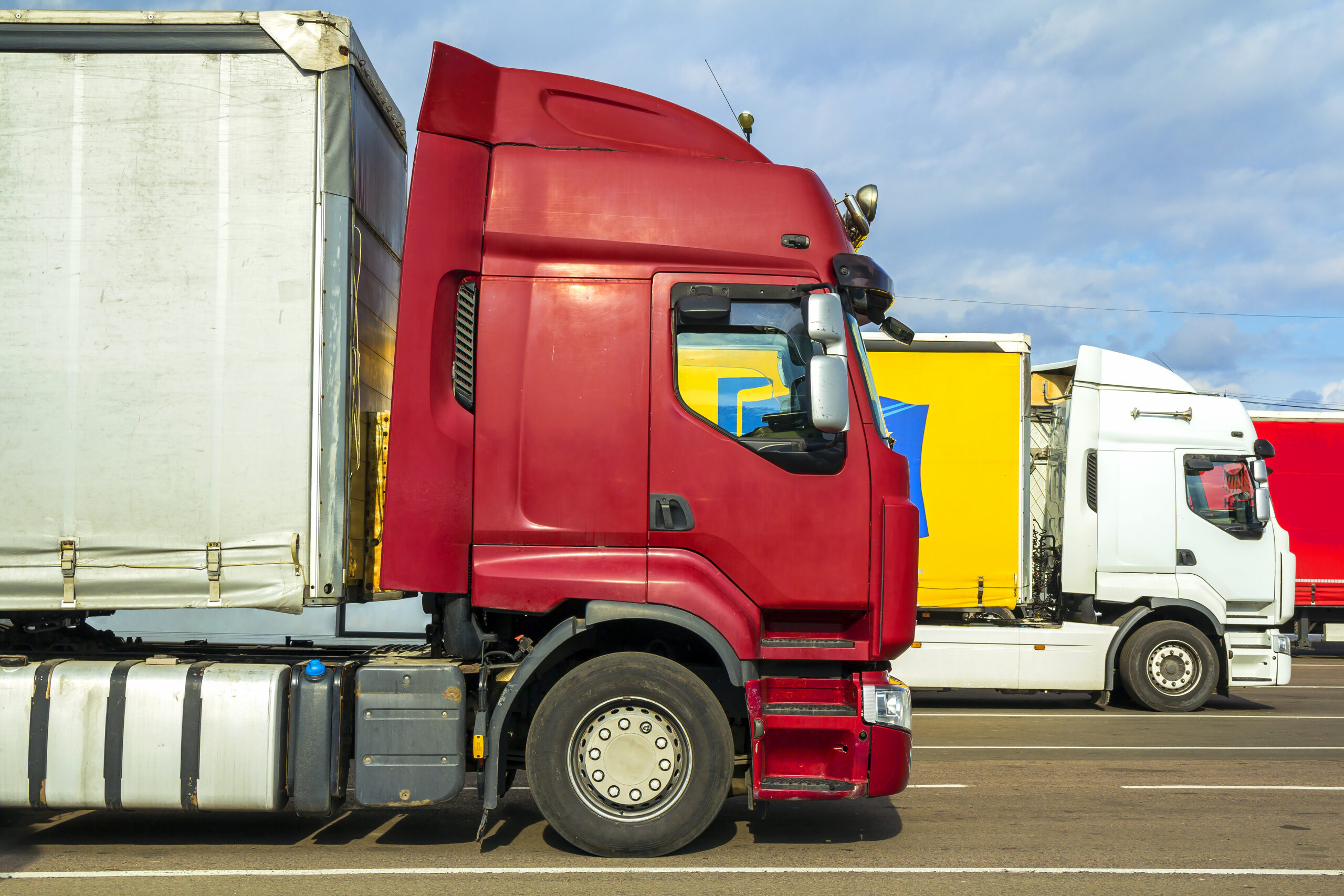 Colorful modern big semi-trucks and trailers of different makes and models stand in row on flat parking lot of truck stop in sunshine